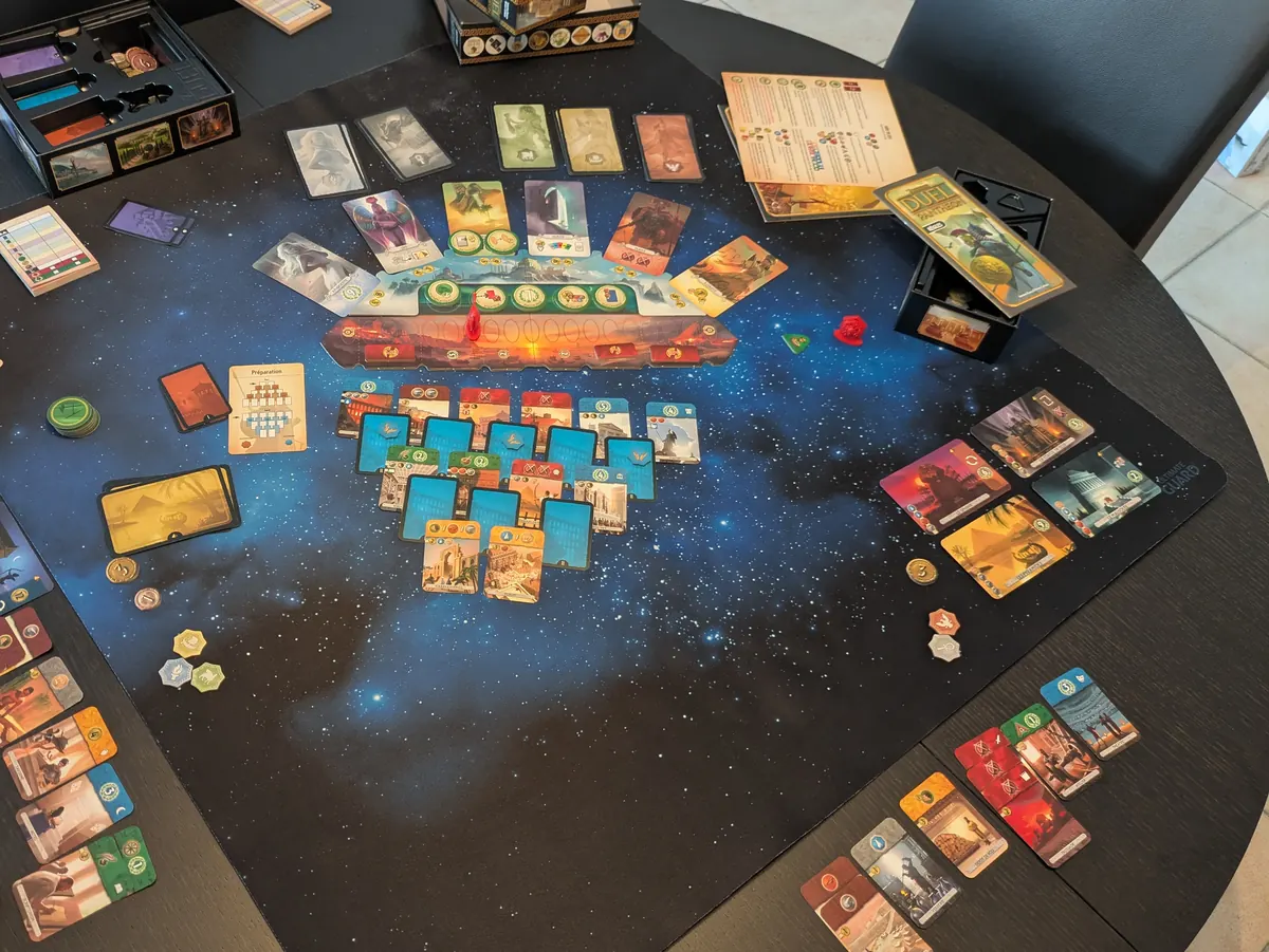 7 Wonders Duel mid-game on a round table: card pyramid, military track and wonder cards spread across a starry playmat