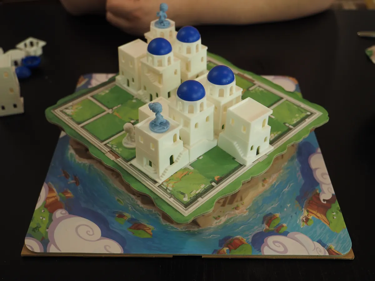 Santorini at the end of a two-player game: white towers with blue domes and worker figures on the island board